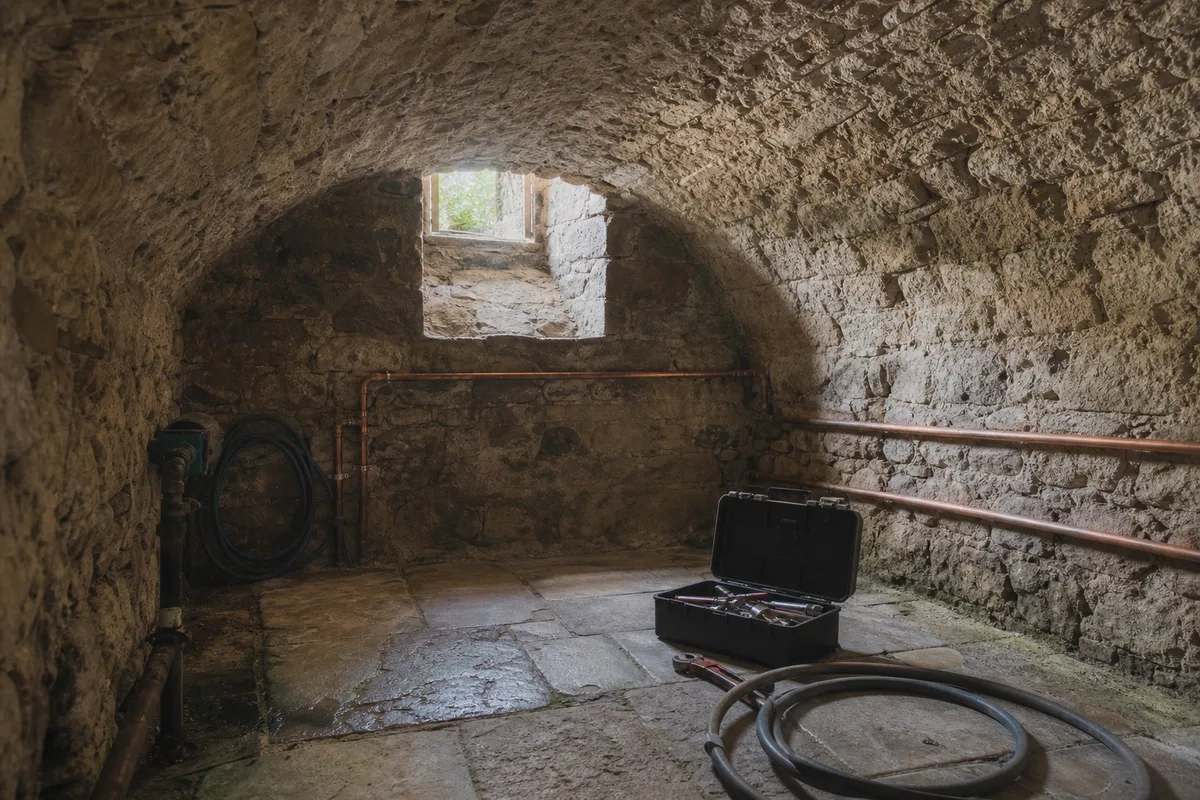 Cave voûtée en pierre avec canalisations apparentes et outillage de plomberie posé au sol, lumière naturelle tamisée